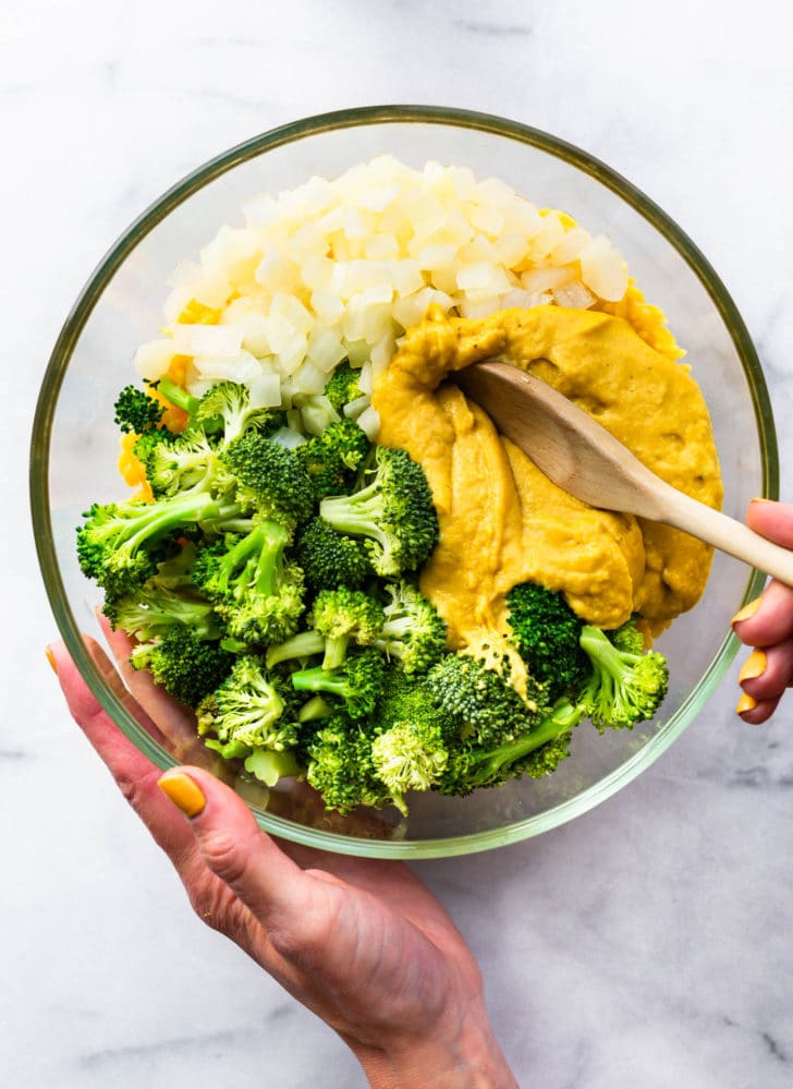 All ingredients for broccoli cheese casserole in clear glass mixing bowl with hand holding wooden spoon ready to stir together.