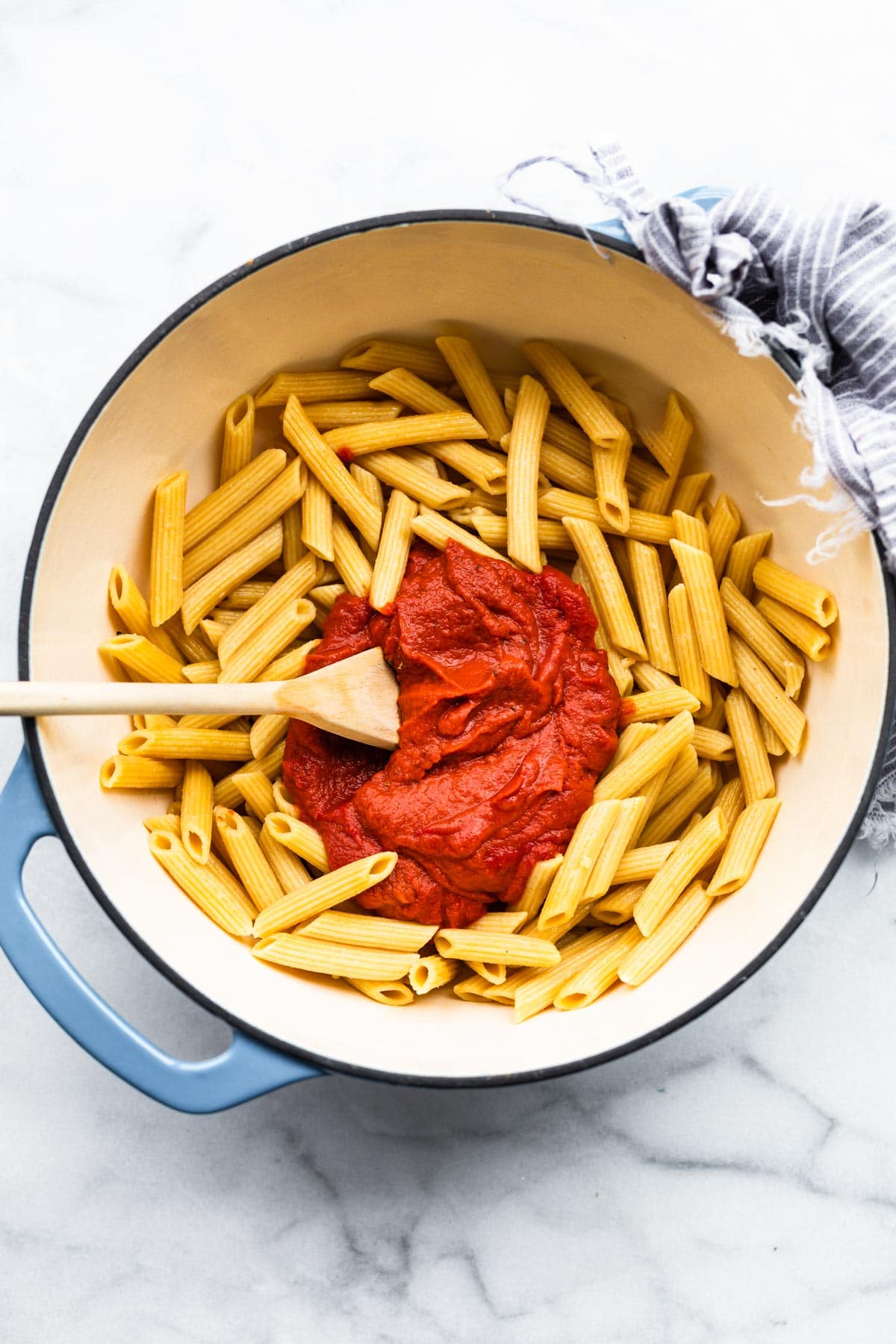 A large pot with a wooden spoon stirring nomato sauce into penne pasta.