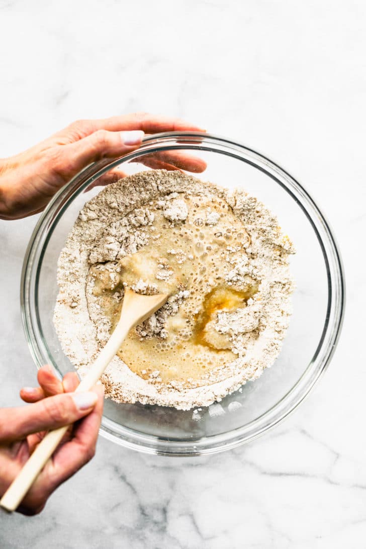 dry ingredients for muffins in a clear bowl being mixed by two hands and a wooden spoon