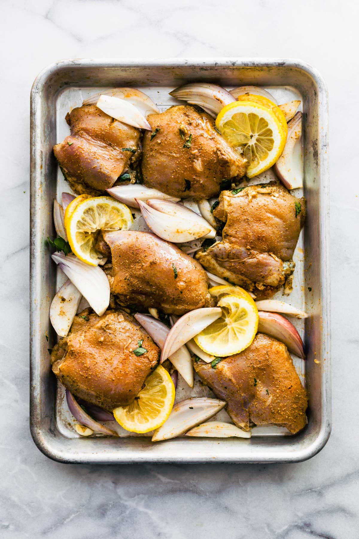 Raw chicken thighs on a sheet pan with slices of lemon and onions.