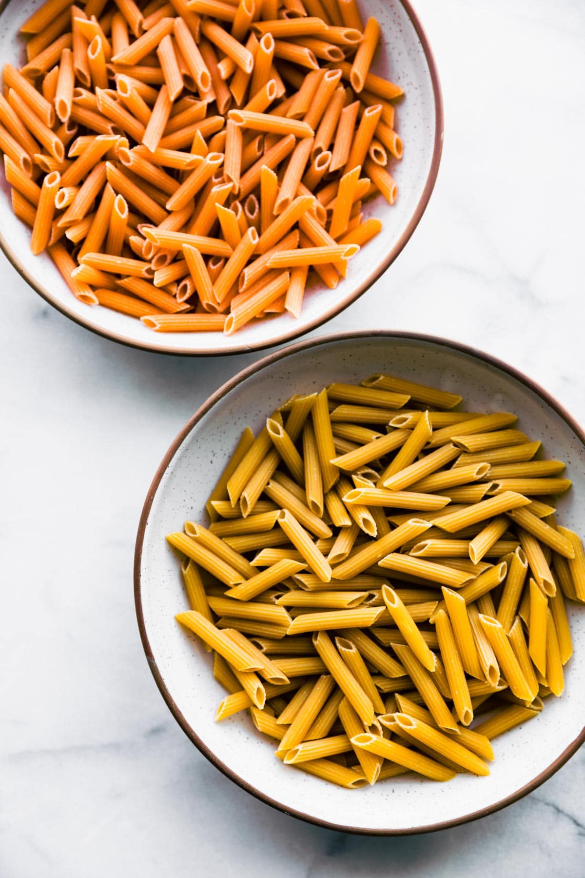 overhead image: 2 bowls of uncooked penne pasta - one with gluten free penne and one with tomato penne