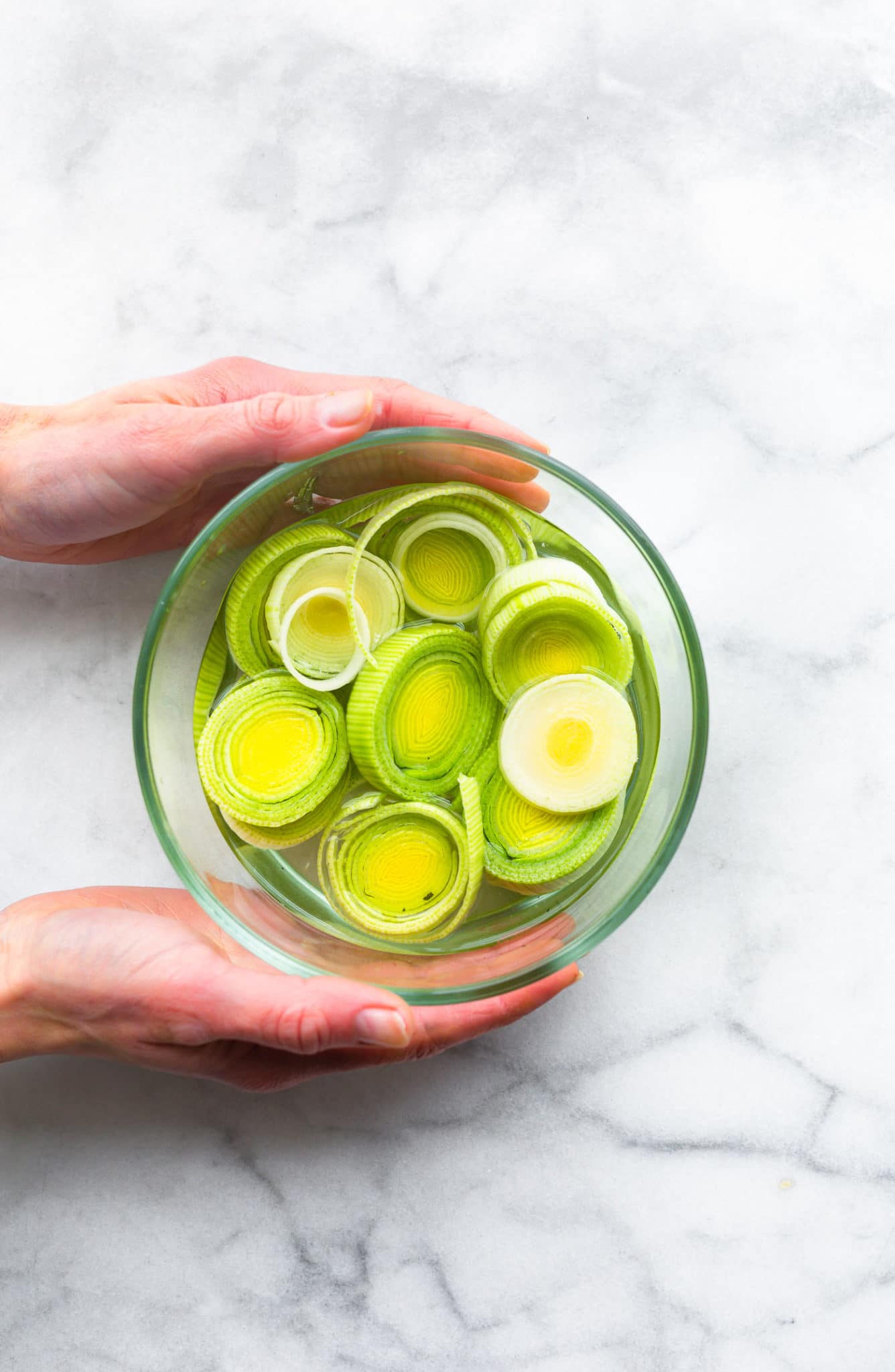 two hands holding a glass bowl full of leeks that have been sliced into rings
