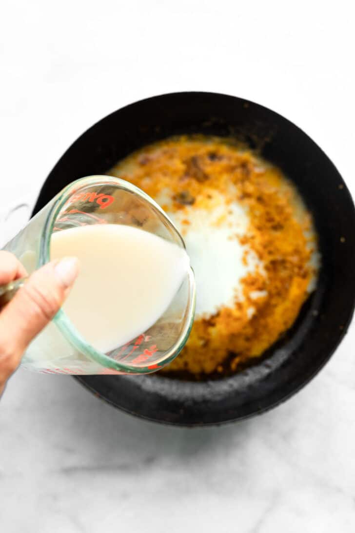 Hand pouring milk from a measuring cup into a skillet to create sauce base.