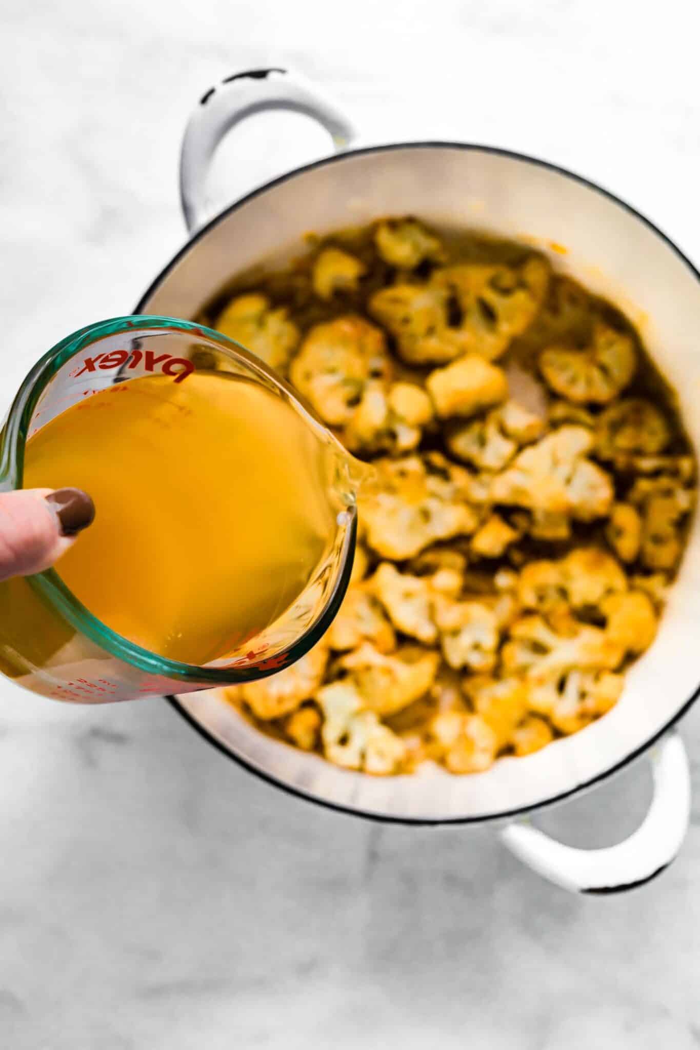 Vegetable broth being poured into a soup pot with roasted cauliflower.