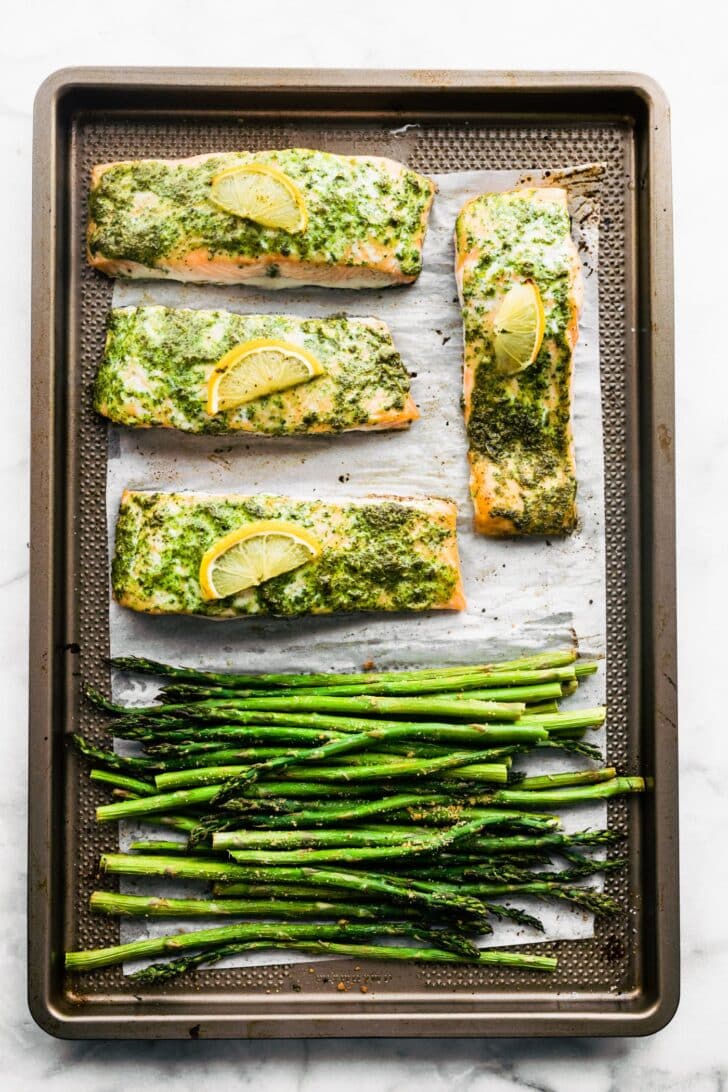 Four salmon filets in herb sauce and lemon slices and asparagus on a sheet pan.