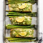 Three salmon filets topped with green herb sauce and lemon slices with asparagus between them.