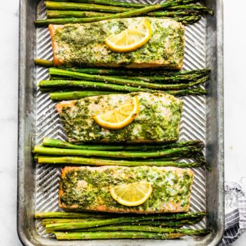 Three salmon filets topped with green herb sauce and lemon slices with asparagus between them.