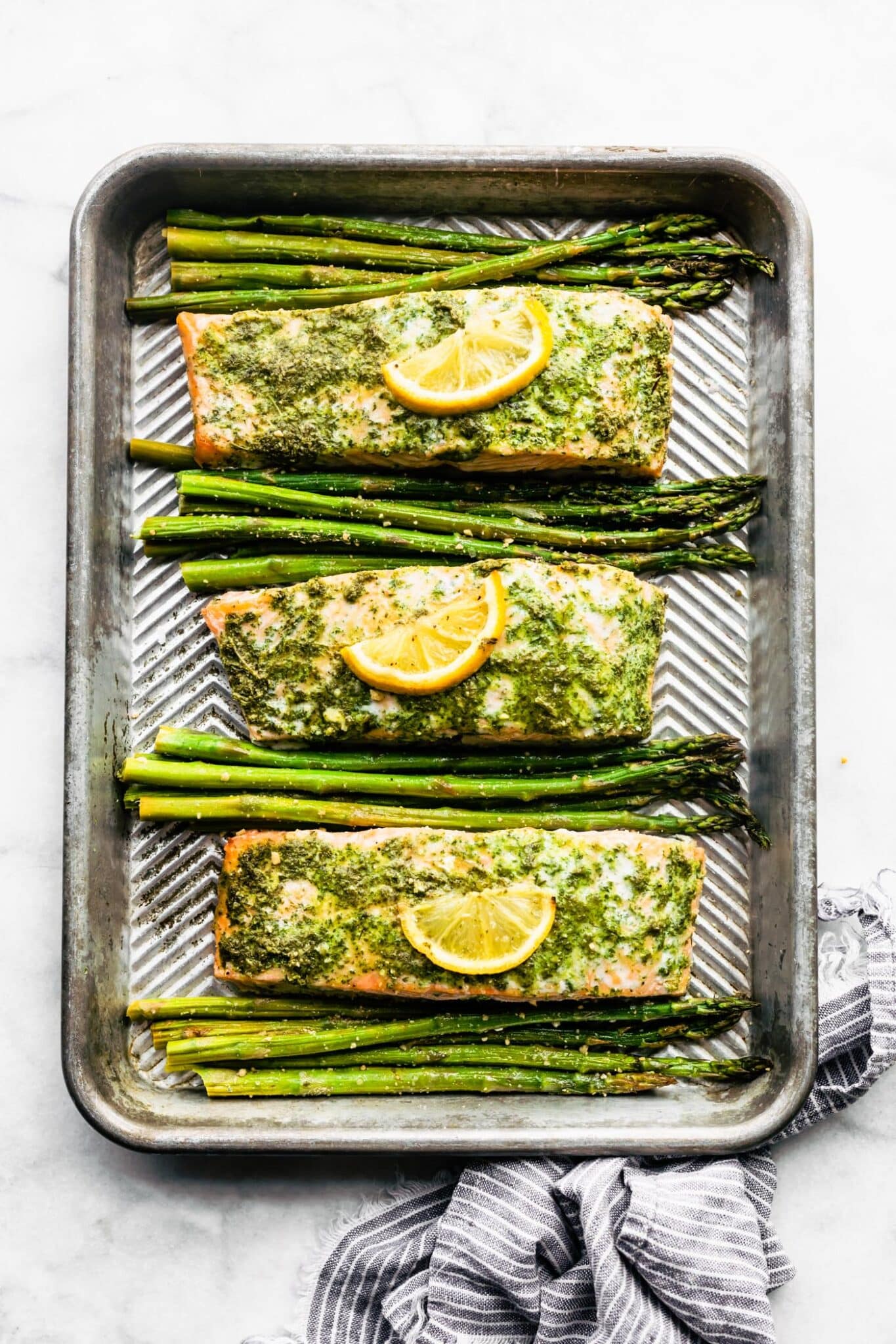 Three salmon filets topped with green herb sauce and lemon slices with asparagus between them.