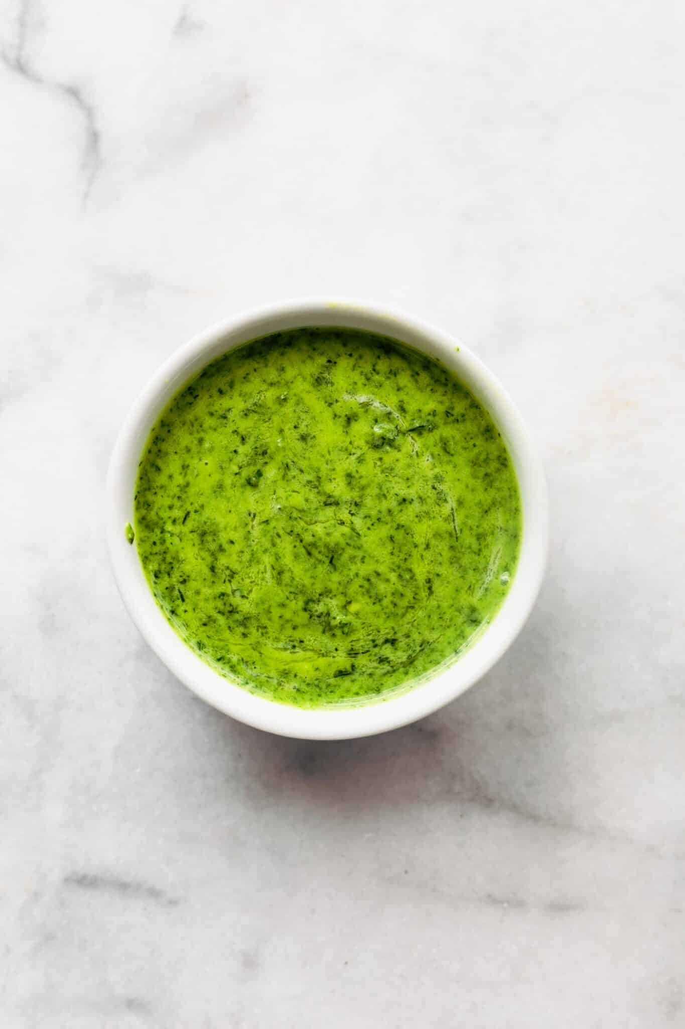 Bright green herb sauce in a small white bowl on a marble countertop.