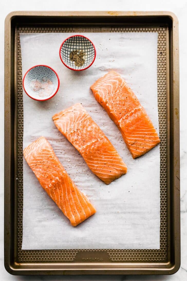 Baking sheet arranged with raw salmon fillets and seasoning bowls