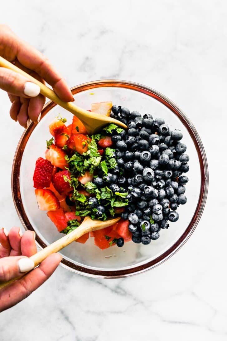 Two hands using wooden spoons to toss summer berry salad in a bowl.