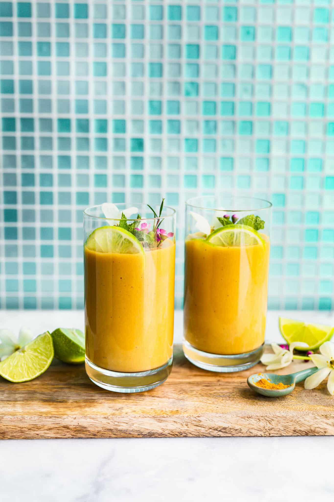 A side shot of two turmeric smoothies in glasses on a serving tray with a garnish of lime, mint, and flowers.