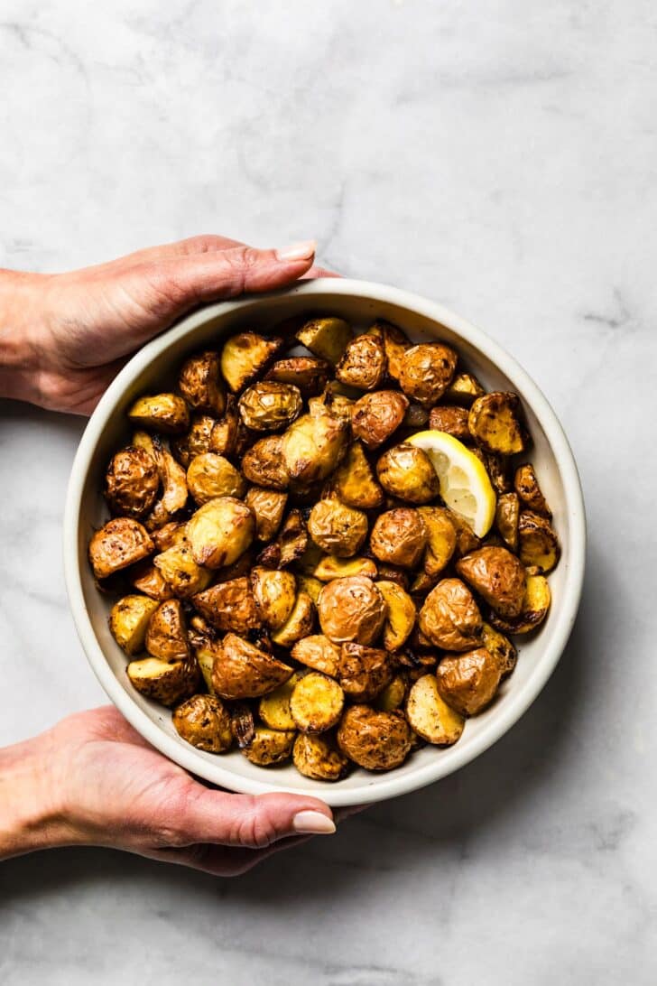 Hands holding a bowl of crispy air-fried baby potatoes with a lemon slice.