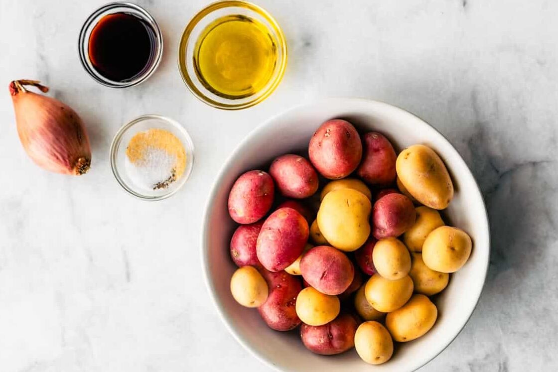 Red and gold baby potatoes in a white bowl, shown alongside small bowls of oil and seasoning, with a shallot placed next to them.