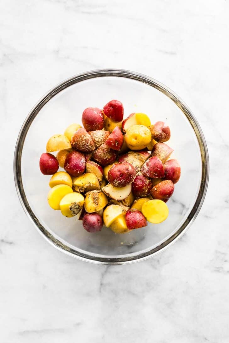 Raw halved baby potatoes in a glass bowl tossed with seasoning.