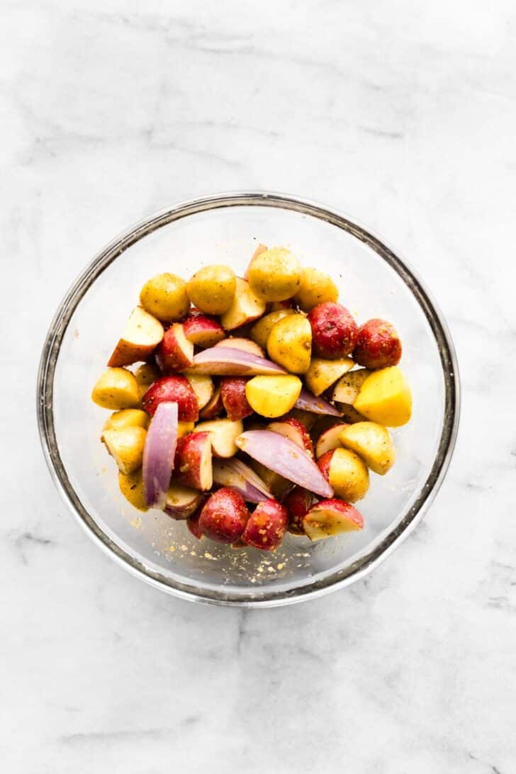 Baby potatoes and sliced shallots mixed with oil and spices in a glass bowl.