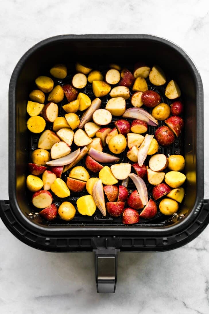 Seasoned baby potatoes and sliced shallots arranged in an air fryer basket before cooking.