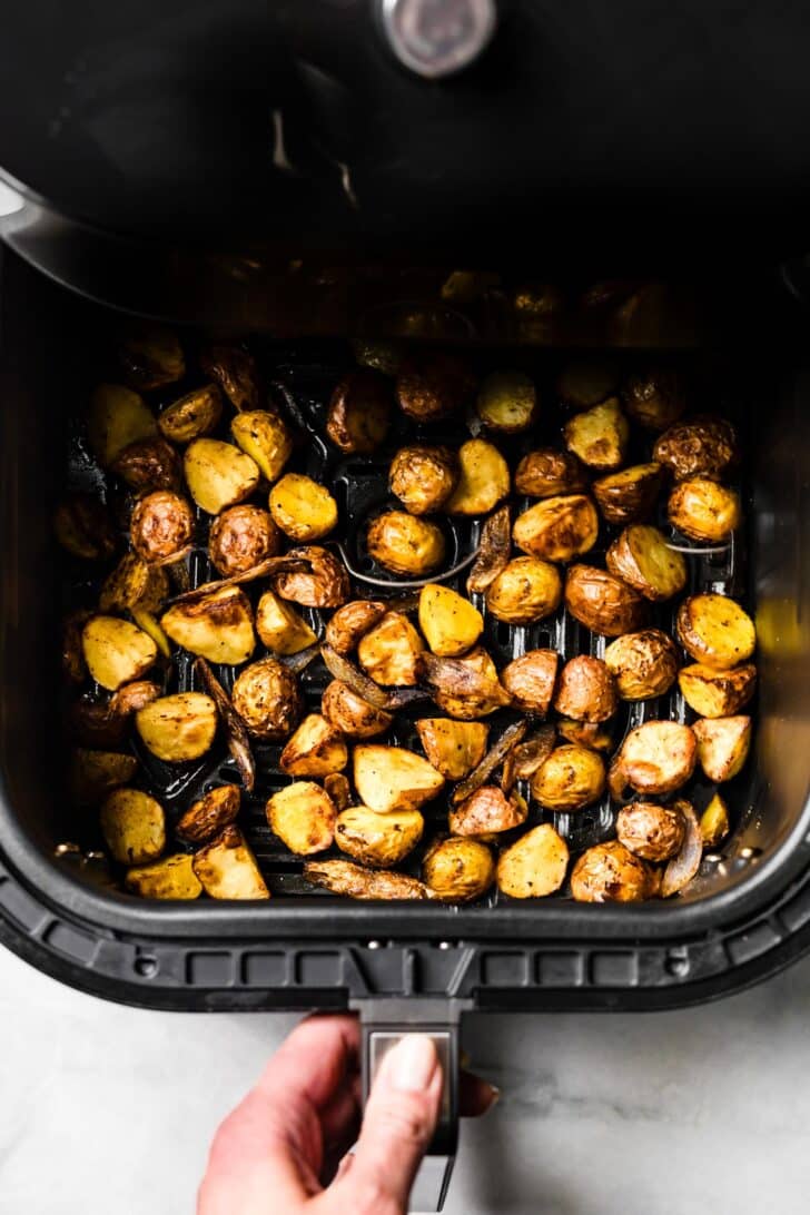 woman opening an air fryer basket filled with golden, crisp baby potatoes and roasted shallots.
