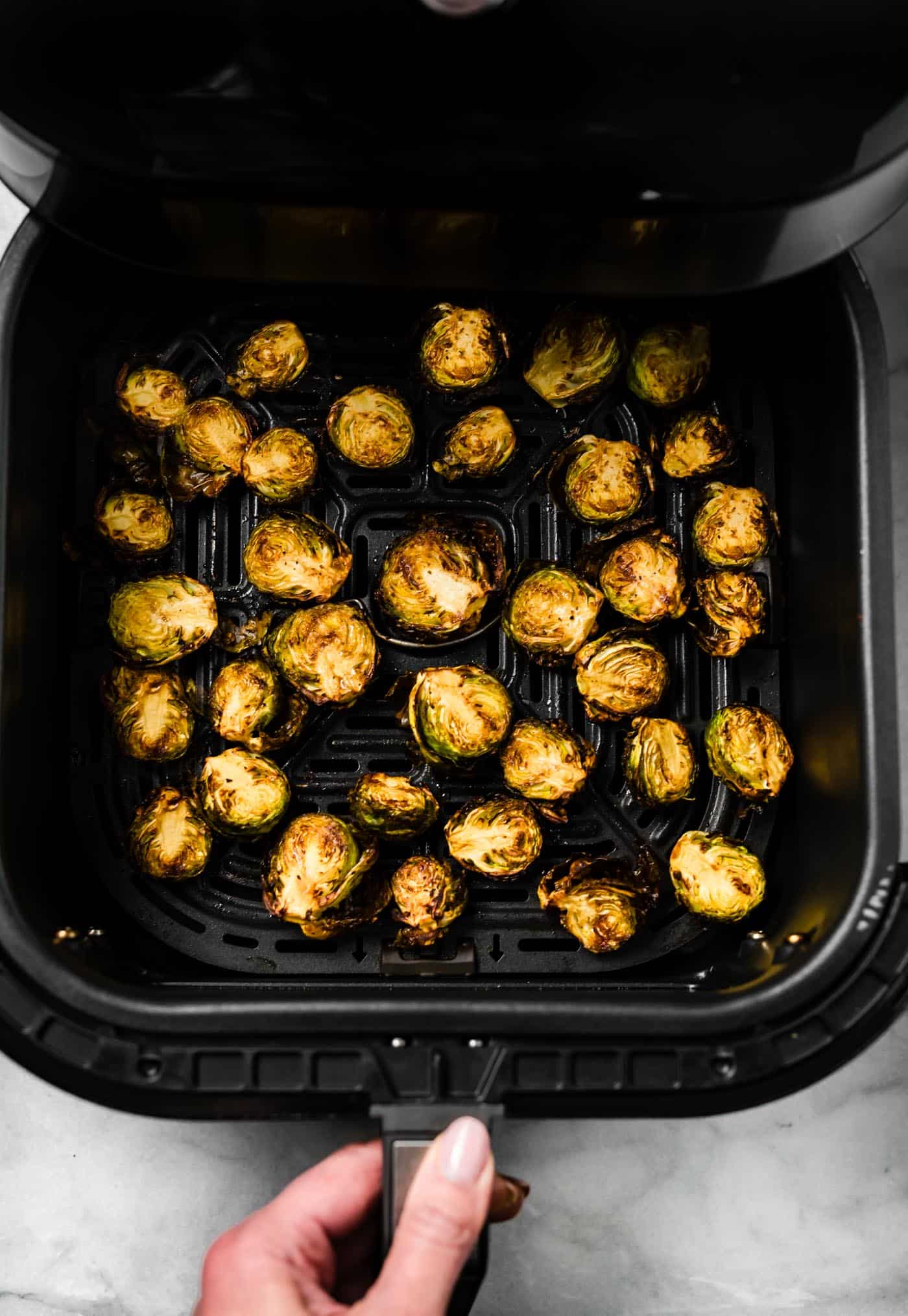 woman's hands opening the air fryer basket full of crispy Brussels sprouts
