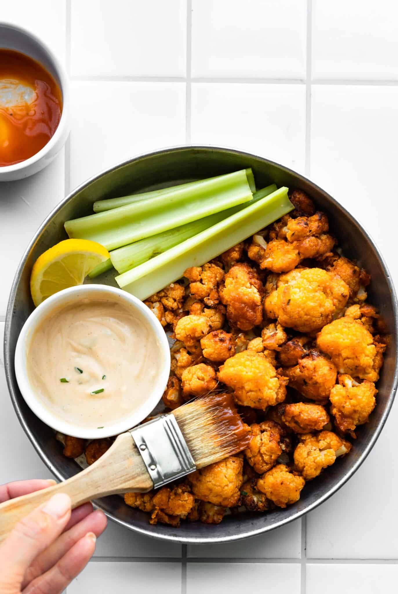 Brushing buffalo sauce on baked buffalo cauliflower in serving dish.