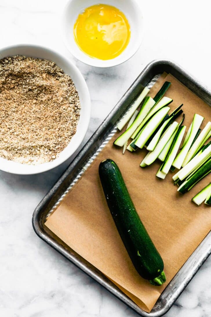 A dredging station for air fryer zucchini fries with a zucchini cut into strips and a whole zucchini.