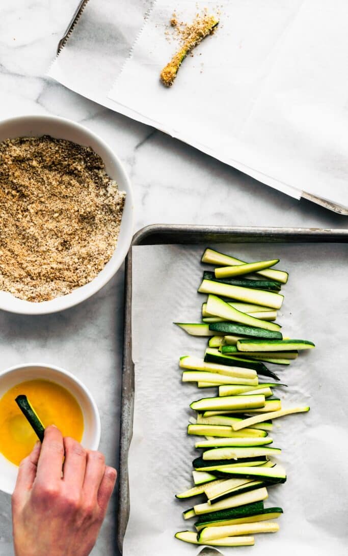 A hand dredging zucchini fries in egg wash and gluten-free breadcrumbs.