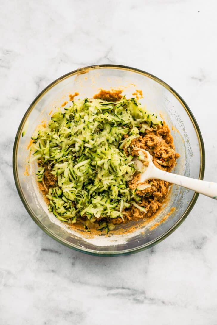 zucchini bread batter in a clear bowl with grated zucchini on top and spatula in the batter.