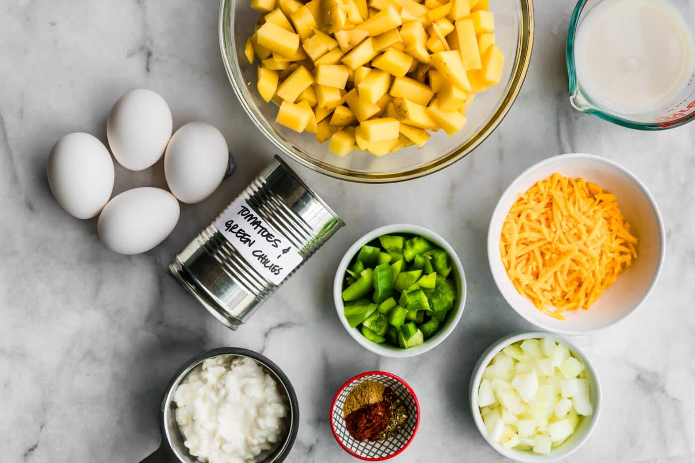Overhead shot of casserole ingredients arranged on a marble surface, including diced potatoes, chopped onion and bell pepper, shredded cheese, cottage cheese, eggs, milk, a can of tomatoes with green chiles, and spices in small bowls.