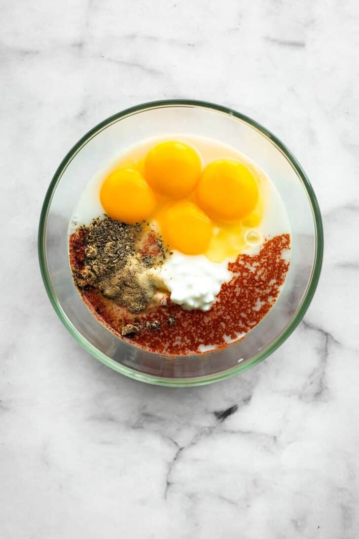 Glass bowl showing eggs, cottage cheese, milk, and colorful seasonings like paprika, oregano, and cumin before whisking together.