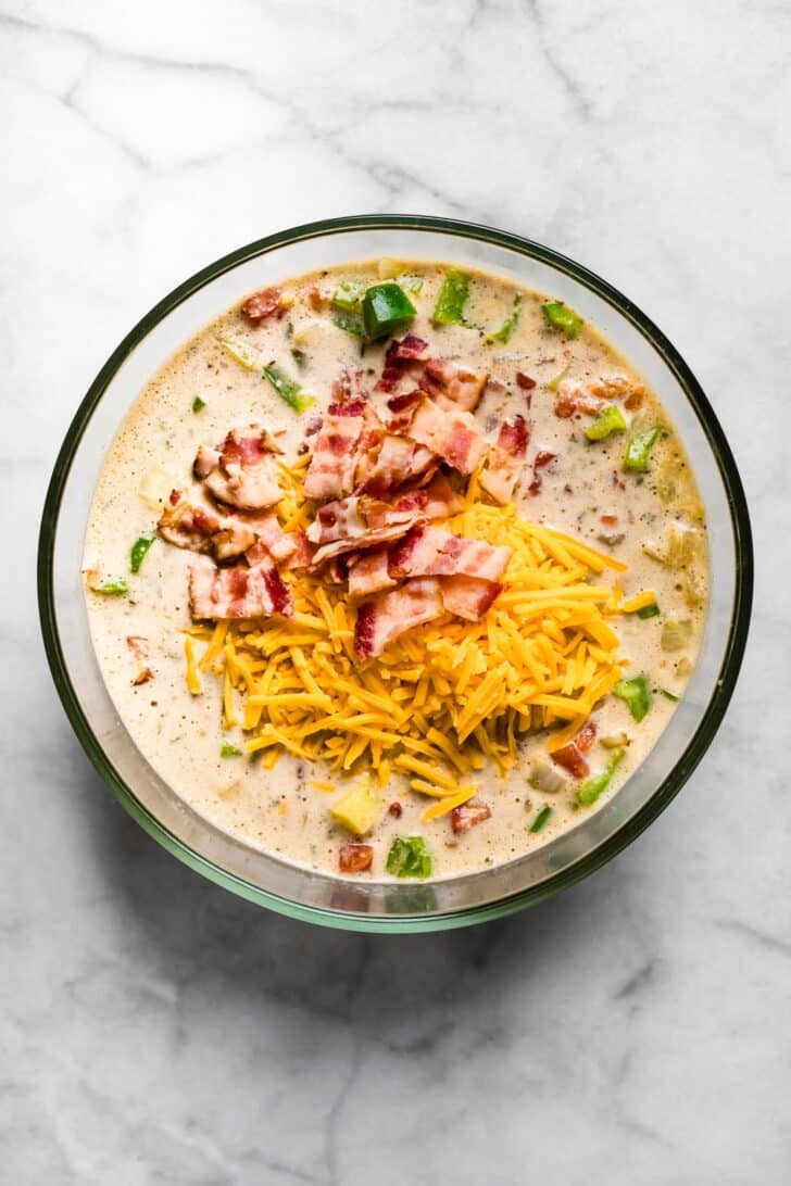 A glass bowl filled with the egg and milk mixture combined with sautéed vegetables, topped with shredded cheese and chopped cooked bacon.