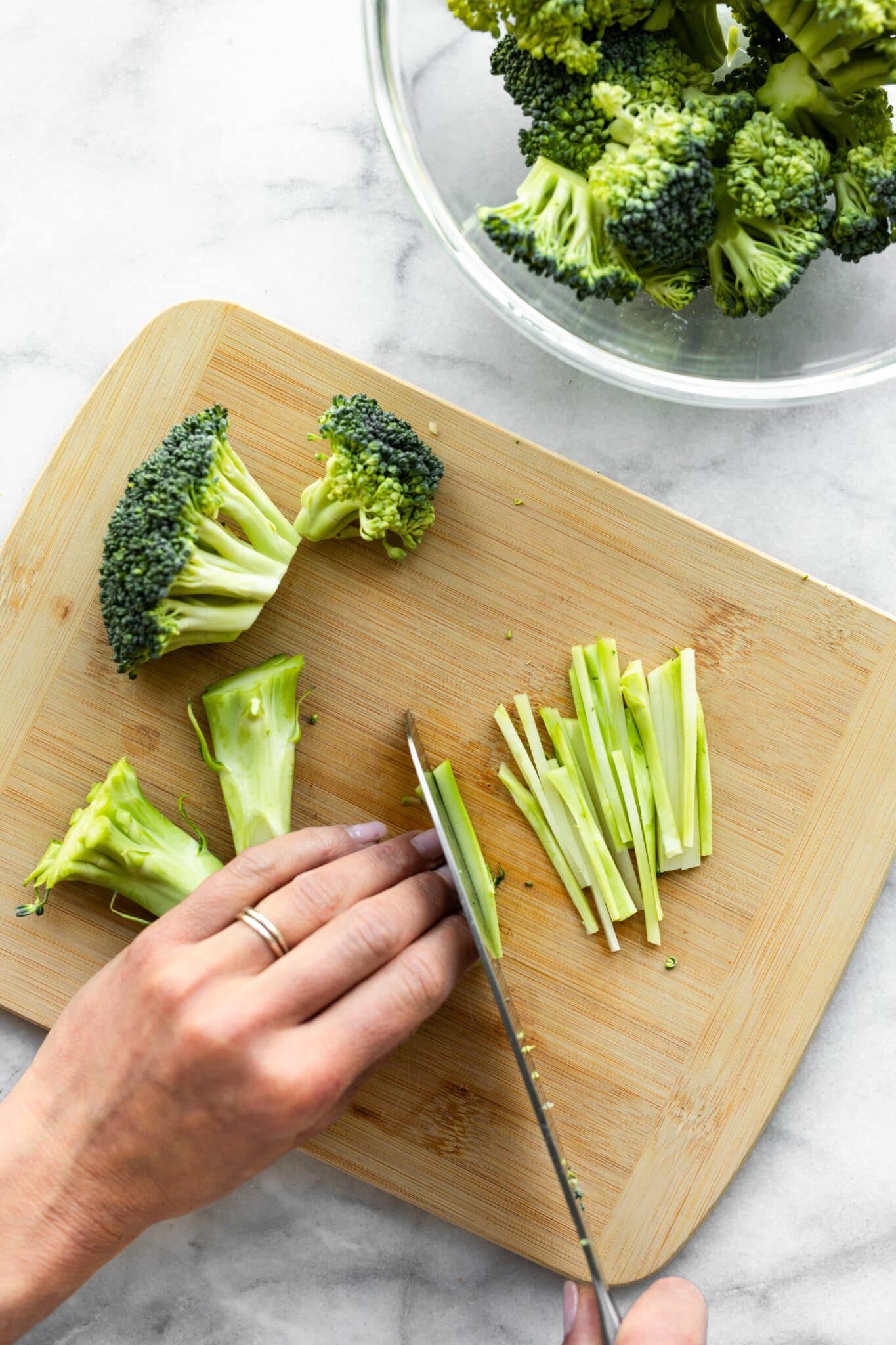 A woman using a knife to thinly slice broccoli stalks on a cutting board.