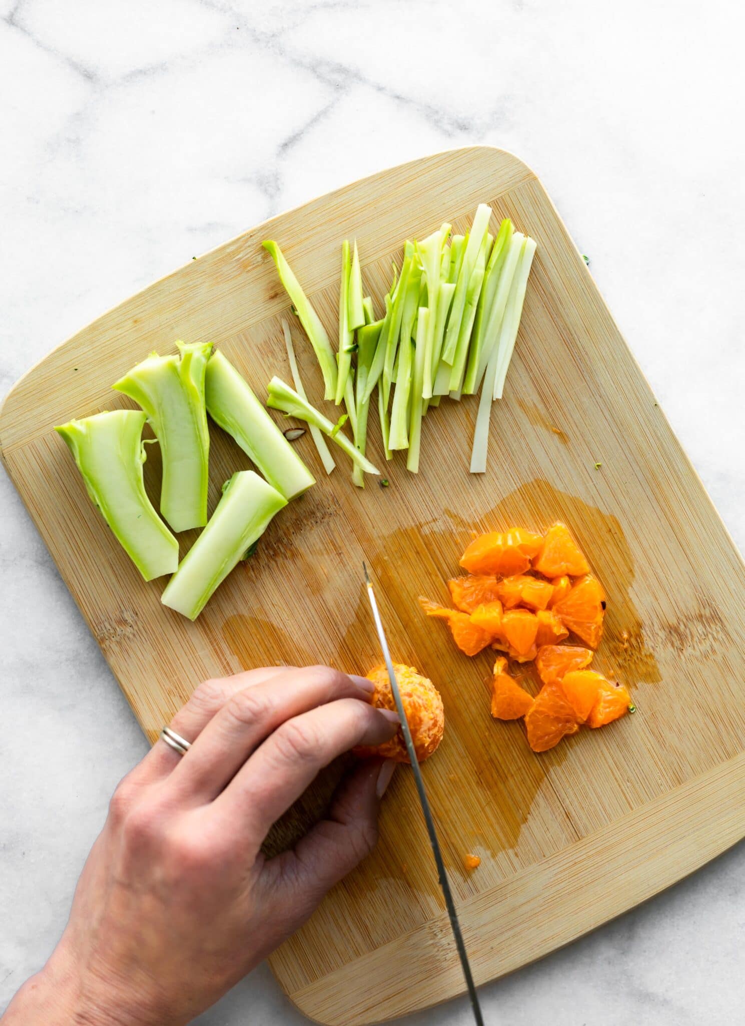 A woman using a knife to thinly slice broccoli stalks and oranges on a cutting board.