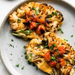 Two roasted cauliflower steaks on a light blue plate with blistered tomatoes and chopped herbs.