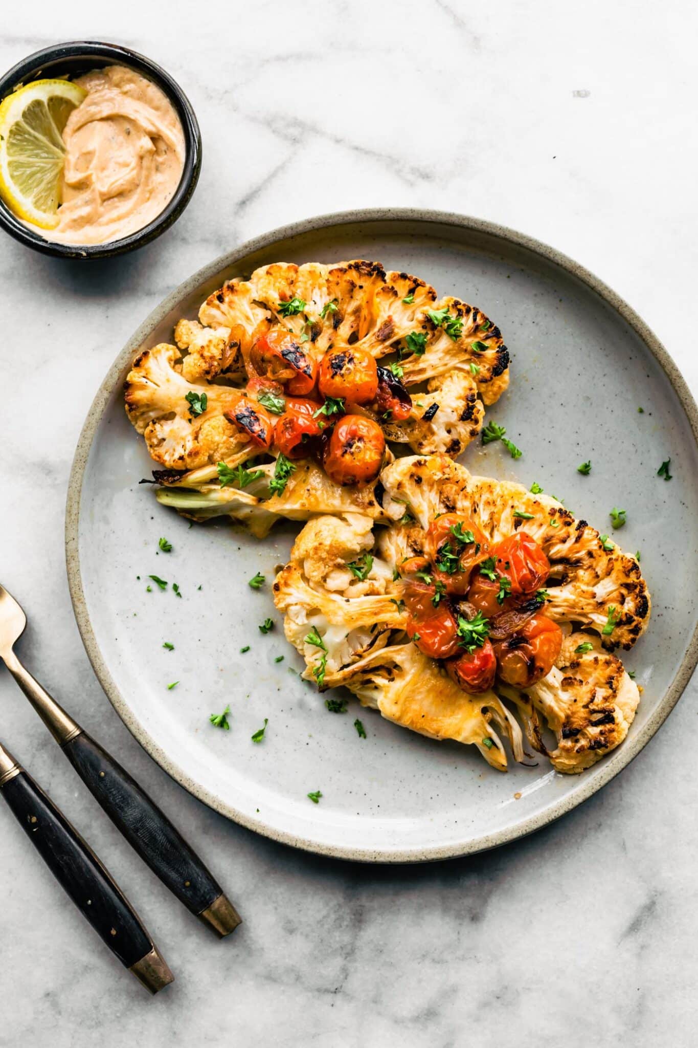 Two roasted cauliflower steaks on a light blue plate with blistered tomatoes and chopped herbs.