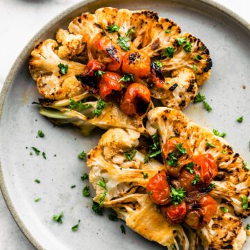 Two roasted cauliflower steaks on a light blue plate with blistered tomatoes and chopped herbs.