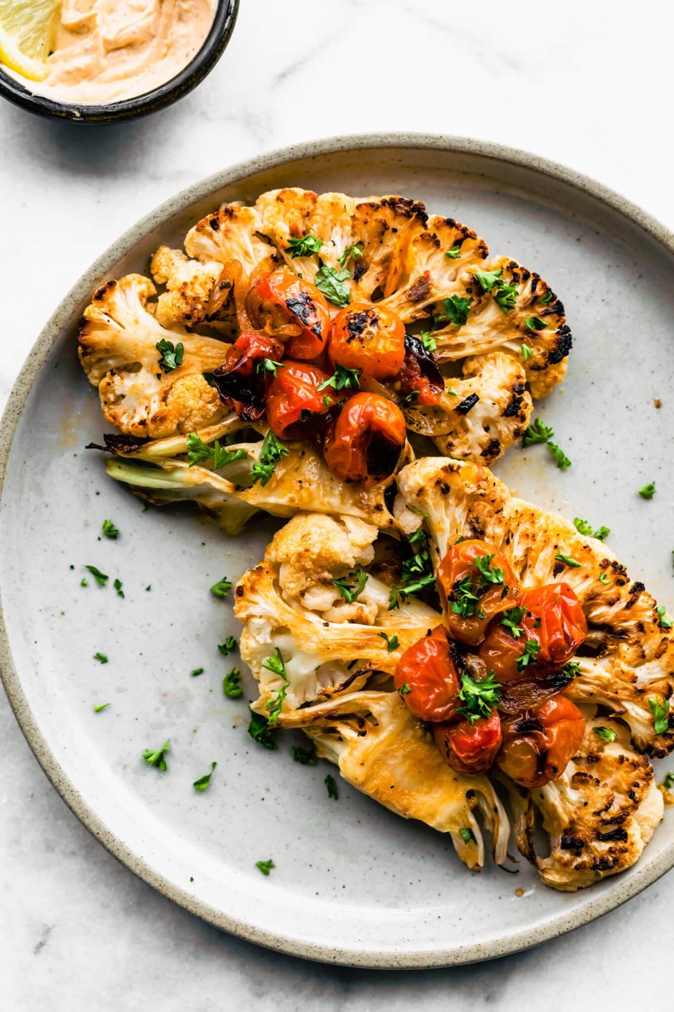 Two roasted cauliflower steaks on a light blue plate with blistered tomatoes and chopped herbs.