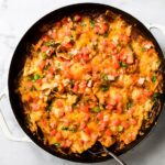 Skillet of baked chicken fajita casserole topped with melted cheese, fresh diced tomatoes, and chopped cilantro, with a serving spoon in it