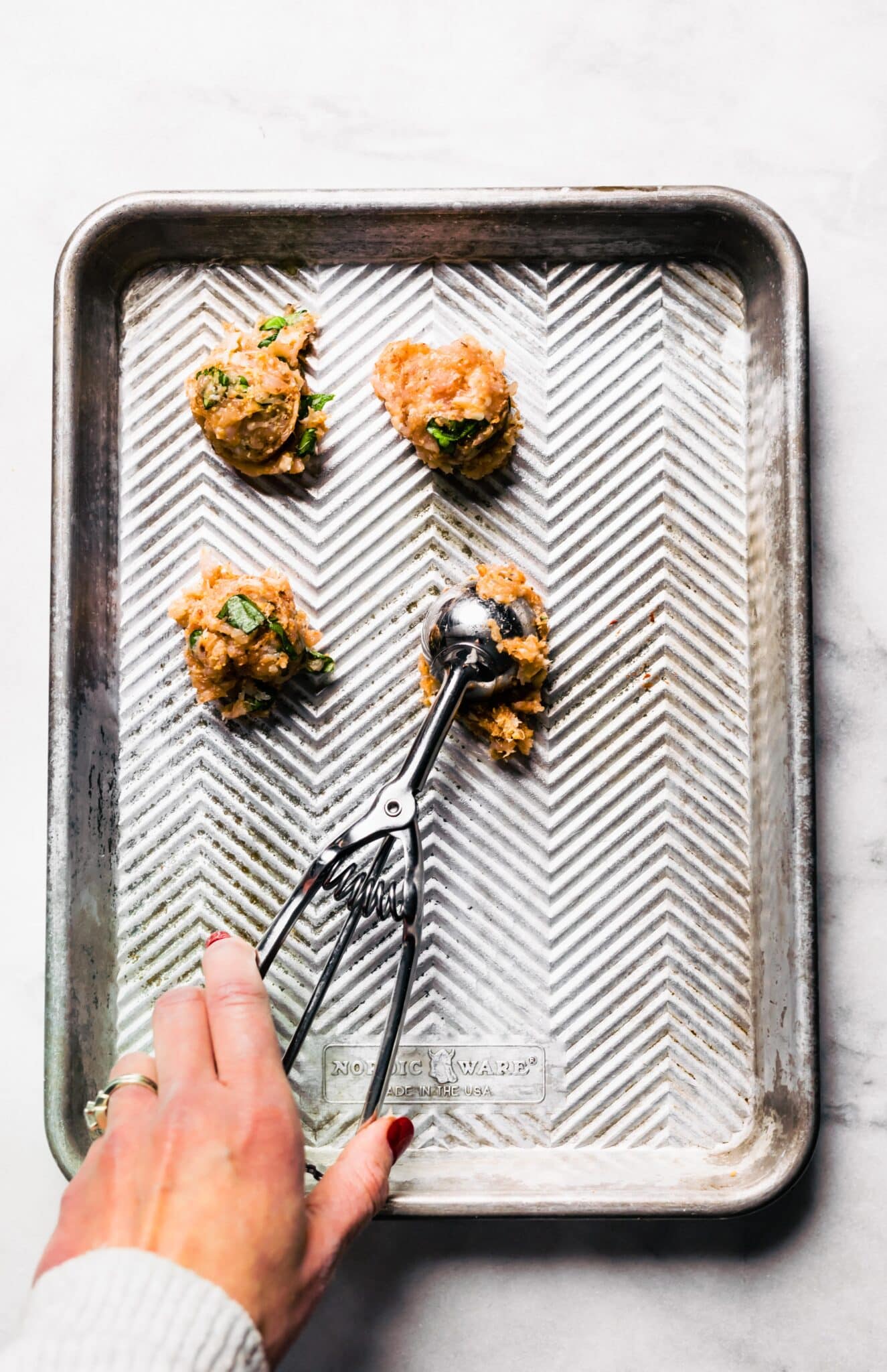 A woman using a dough scoop to spoon meatball batter onto a sheet pan.