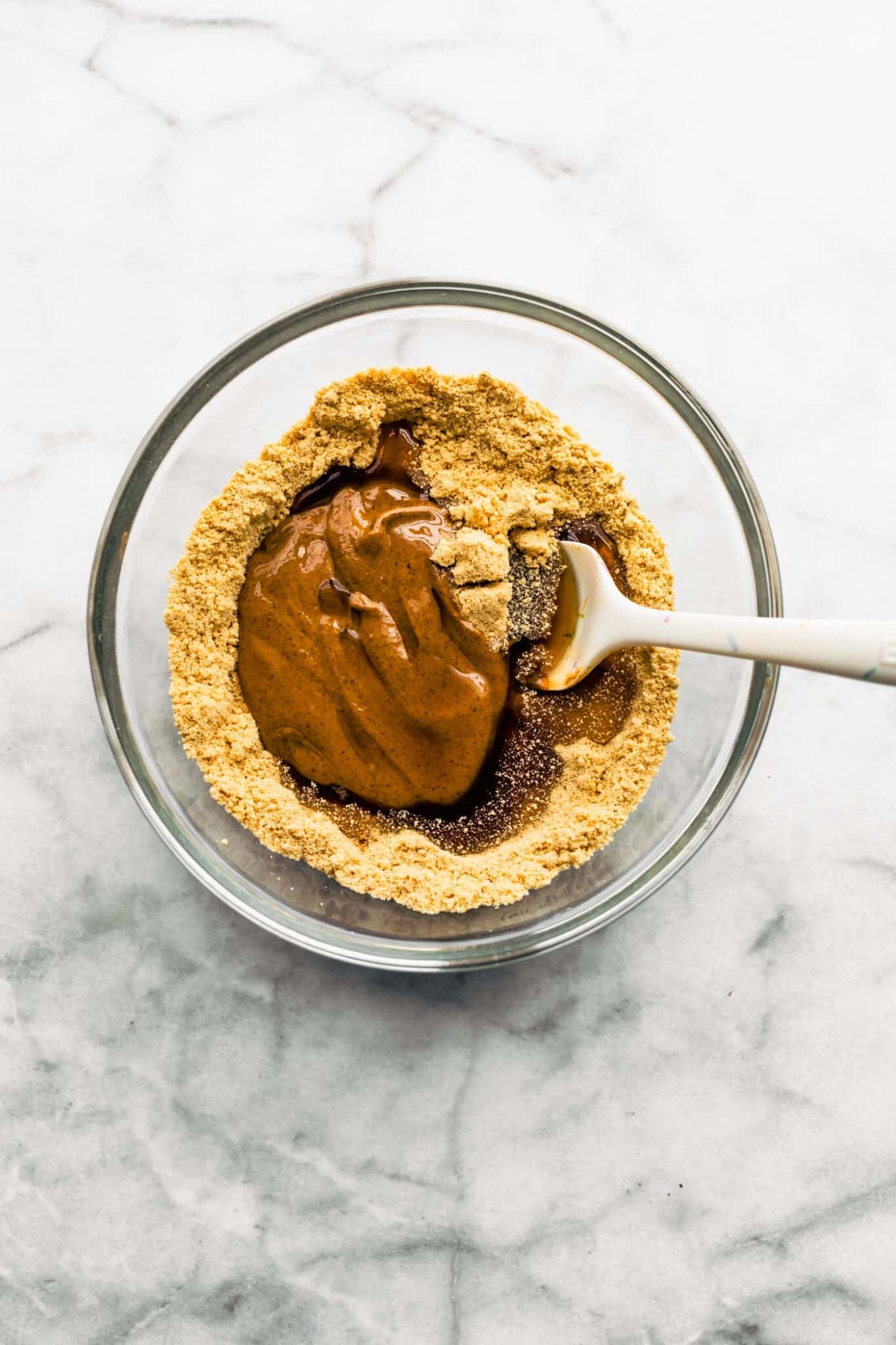 Almond butter and maple syrup in a bowl of dry ingredients with a white spatula.