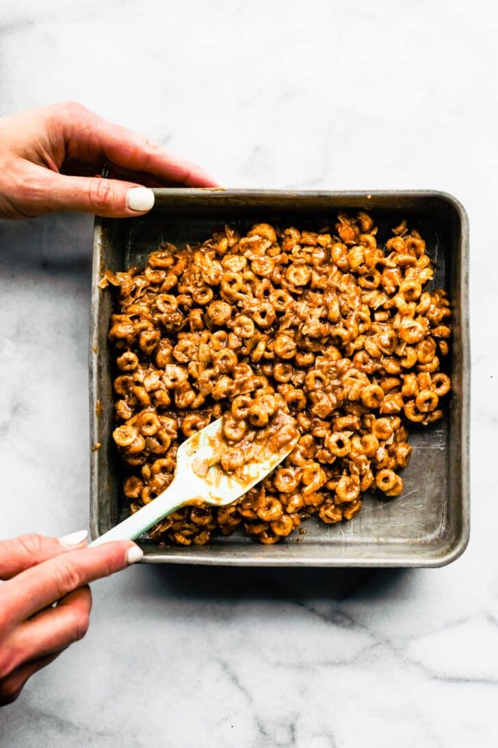A woman's hand using a spatula to press a cereal mixture into a pan.