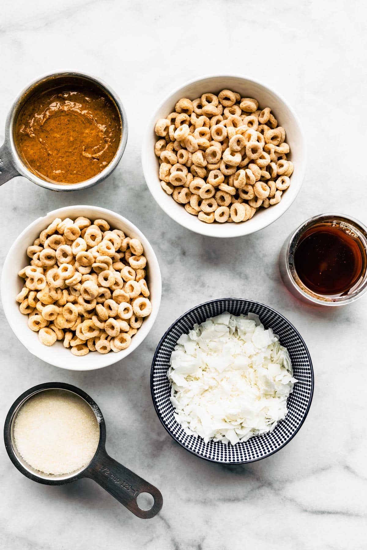 Bowls of puffed cereal, nut butter, shredded coconut, white sugar and honey on a white marble countertop ready to make no bake cereal bars.