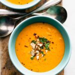 overhead image of a blue bowl filled with root vegetable soup topped with red chili flakes, crushed nuts, and fresh herbs with two spoons on the side