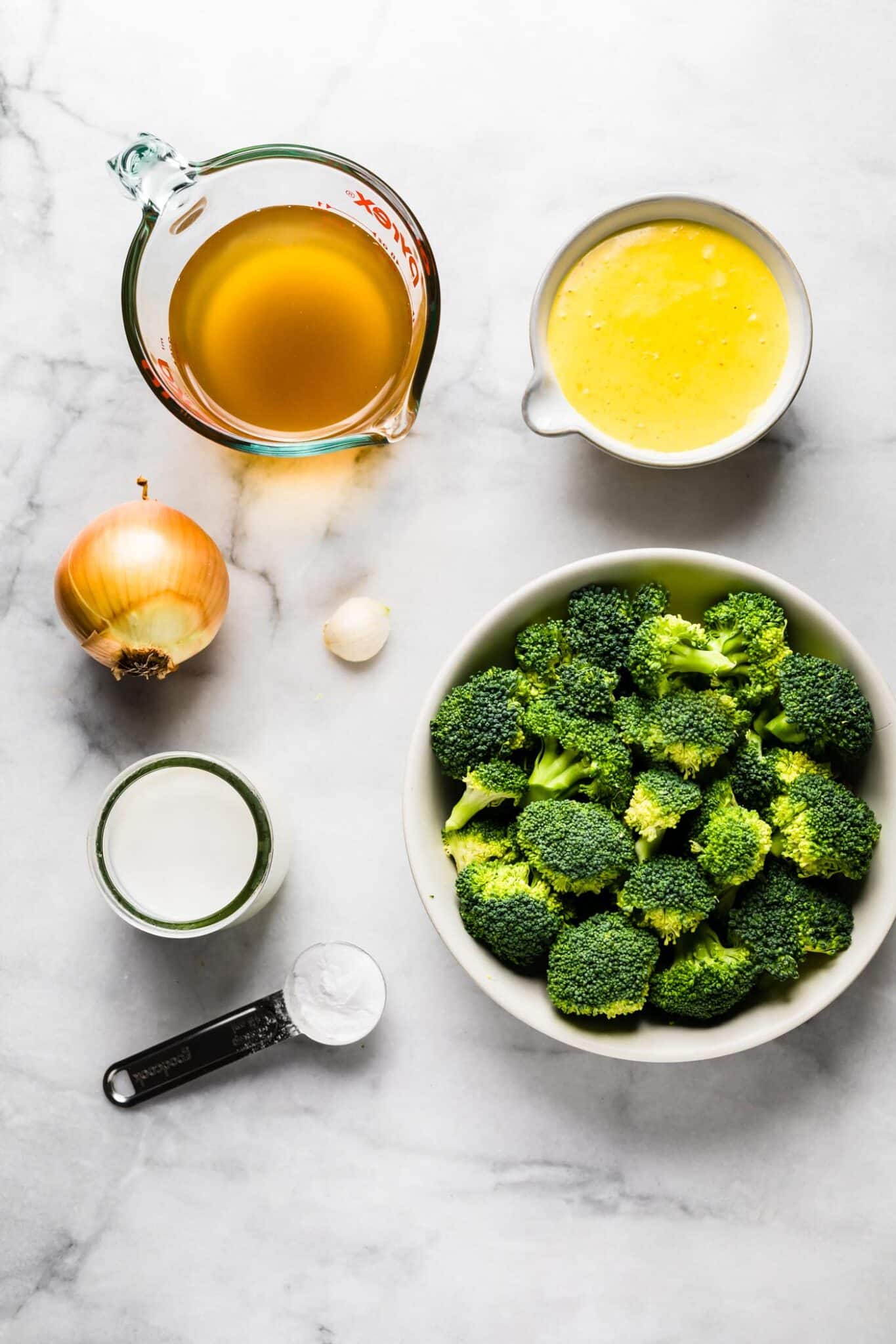 overhead shot of ingredients to make broccoli cheese soup with vegan cheese sauce in a bowl, broccoli floret, broth in a measuring cup, onion, garlic.