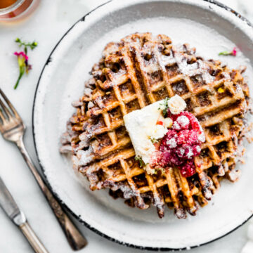 Stack of Paleo waffles on white plate topped with mashed berries, maple syrup, and powdered sugar.