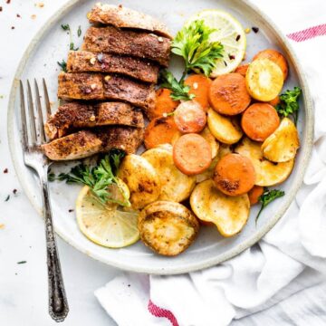 cinnamon chipotle spice rubbed pork chop sliced into pieces on a white plate with roasted carrots and parsnips on the side with a fork and lemons and herbs for garnish