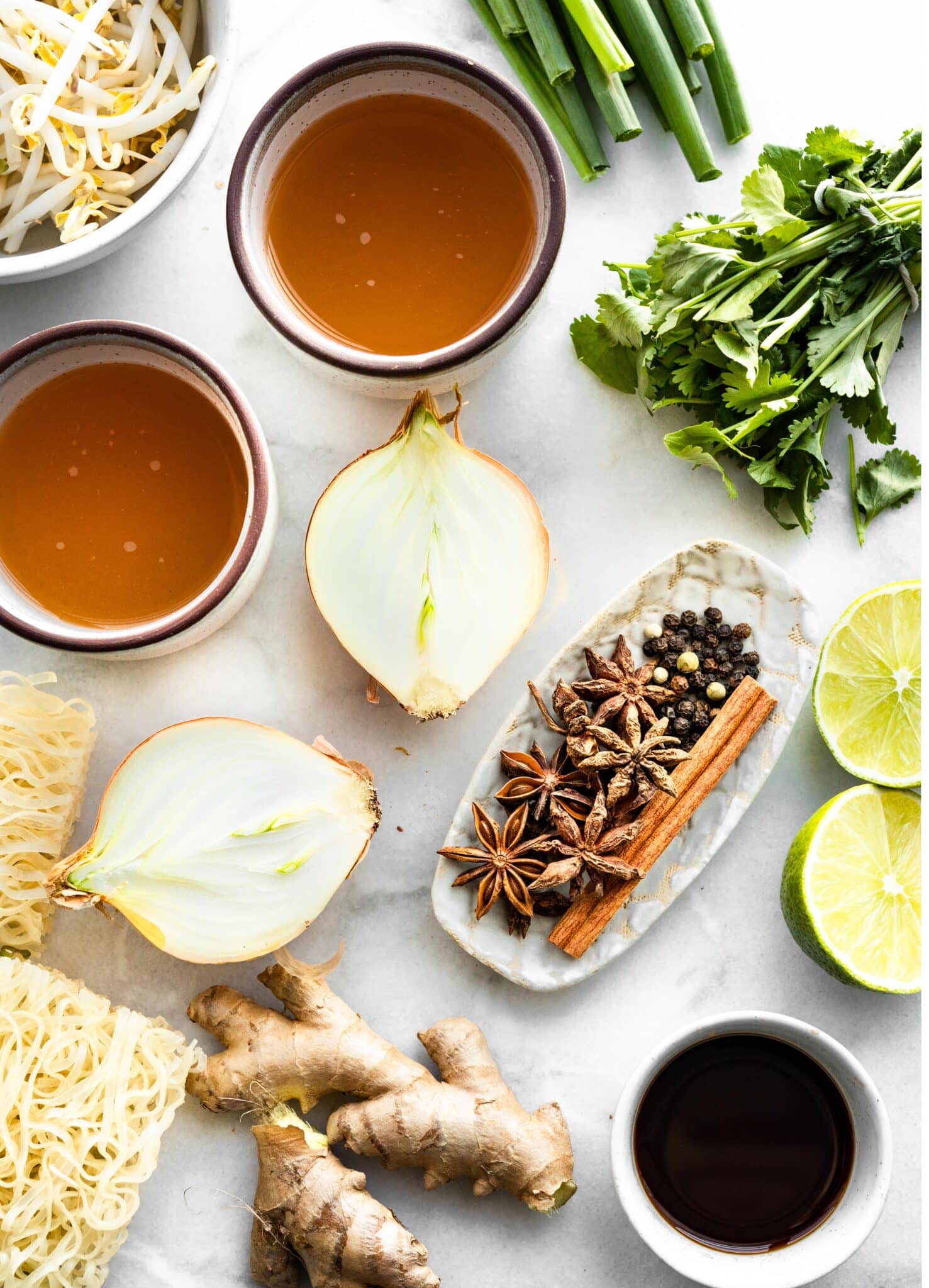 Halved onion, chicken stock, spices, fresh ginger, rice noodles and herbs on a white countertop.