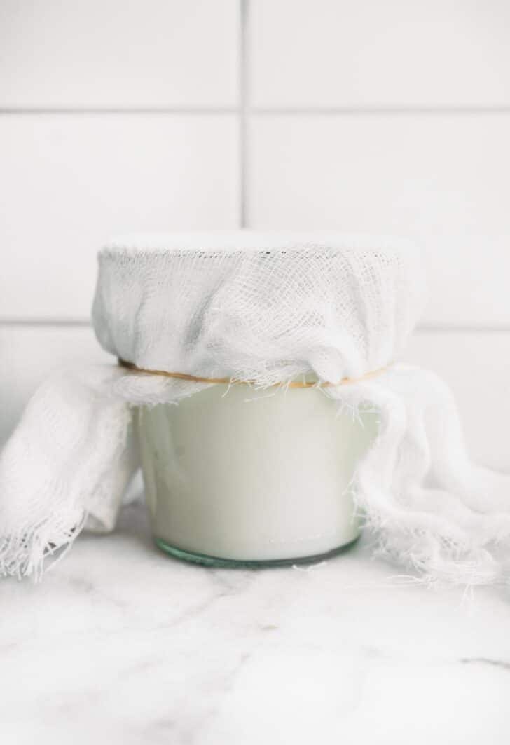 A glass jar full of coconut milk yogurt covered with a loose cloth to ferment.
