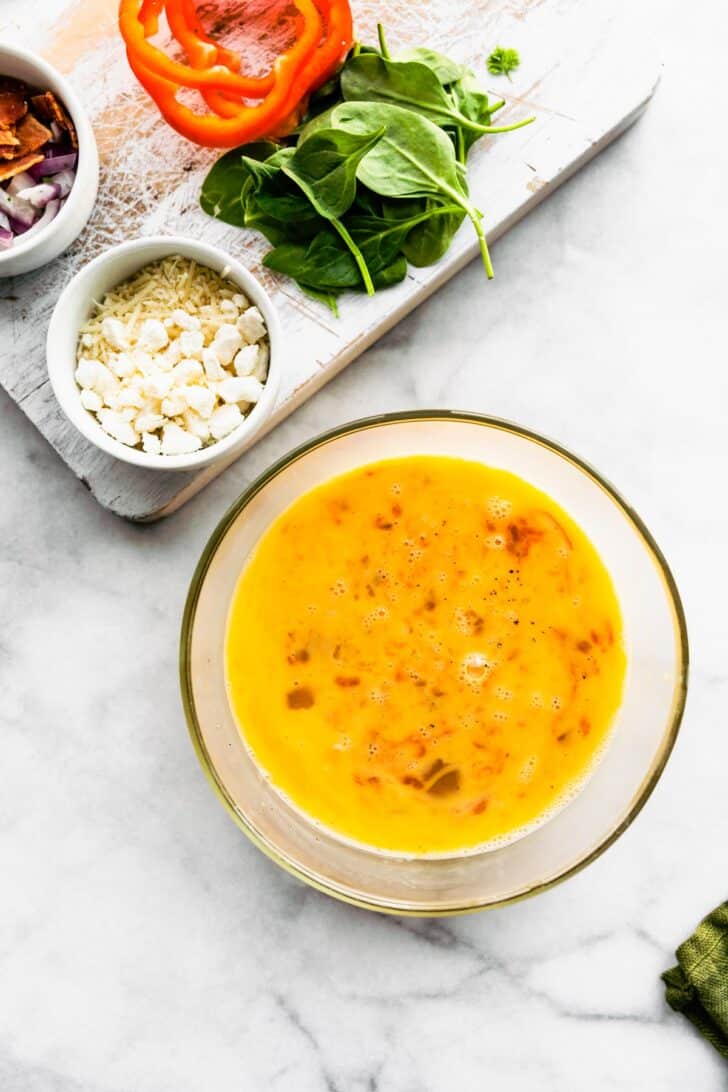 Egg base in a glass bowl and assorted ingredients on a cutting board.