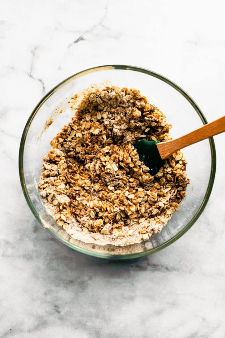 Crumble mixture of oats, flour, melted butter, sugar, and spices in a glass bowl with spatula
