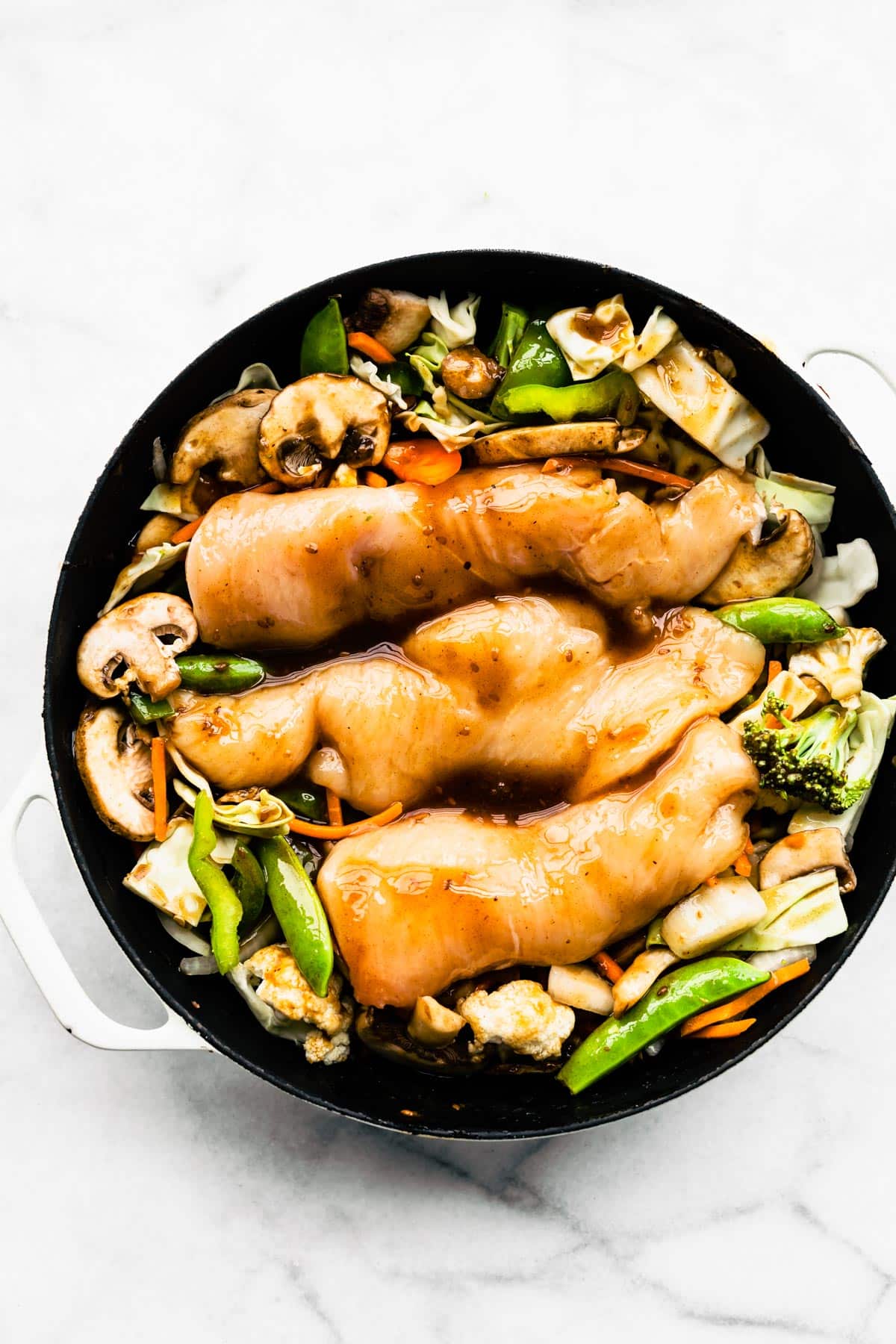 Raw chicken glazed with teriyaki sauce sitting on Asian veggies in an oven safe pan.